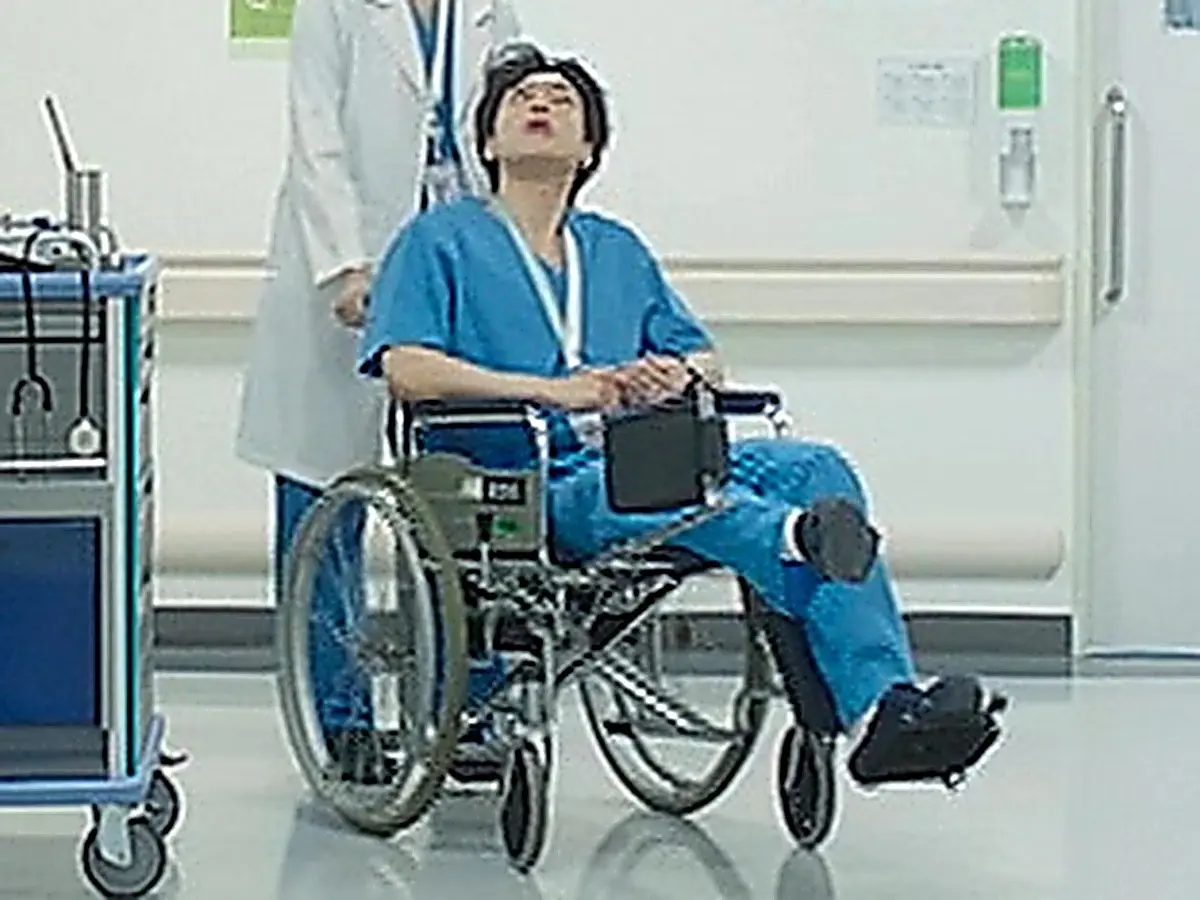 白衣の人物に車椅子を押されながら天井を見上げる青いスクラブ姿の男性